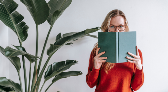 Links im Bild steht eine große Zimmerpflanze. rechts daneben befindet sich eine Frau, die ein grünes Buch so vor sich hält, dass nur der obere Teils des Gesichts zu sehen ist.