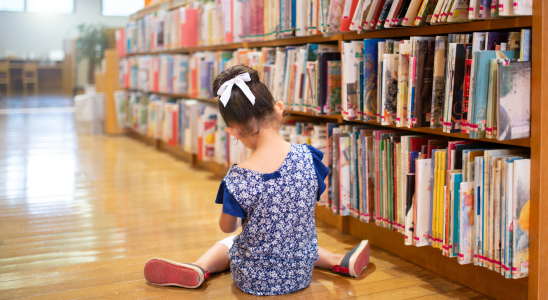 Kind sitzt auf dem Holzboden vor einem gefüllten Bücherregal mit dem Rücken zur Kamera.