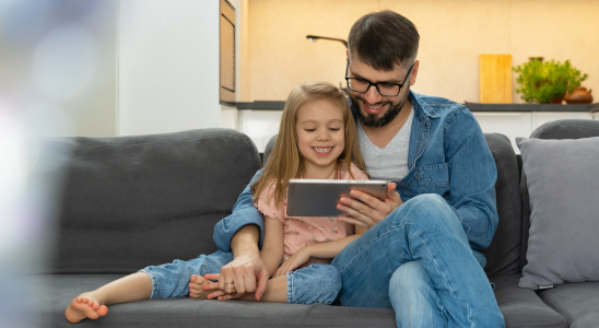 Erwachsene Person und Kind sitzen zusammen auf einer Couch. Erwachsene Person hält ein Tablet in der Hand, das Kind und er gucken zusammen auf das Tablet.