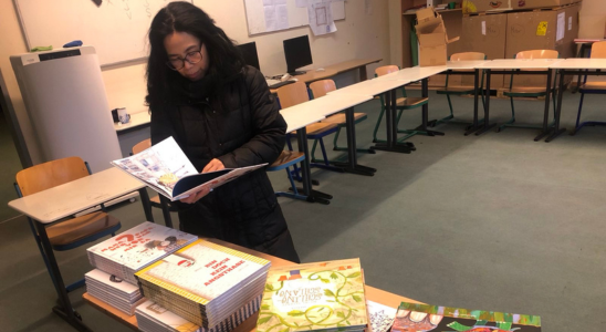 Min Tjoa-Schneider steht in einem großen Raum mit mehreren Tischen. Auf einem Tisch liegen mehrere Stapel Kinderbücher. Min blättert in einem der Bücher.